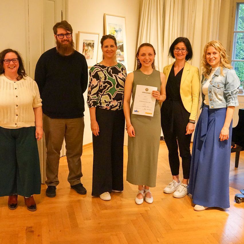 Eine Gruppe von sechs Personen, darunter eine Frau mit einem Zertifikat, posiert für ein Foto in einem Raum. Sie alle lächeln.