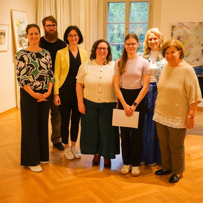 Eine Gruppe von Menschen steht in einer Kunstgalerie und lächelt. Eine Frau hält ein Stück Papier.