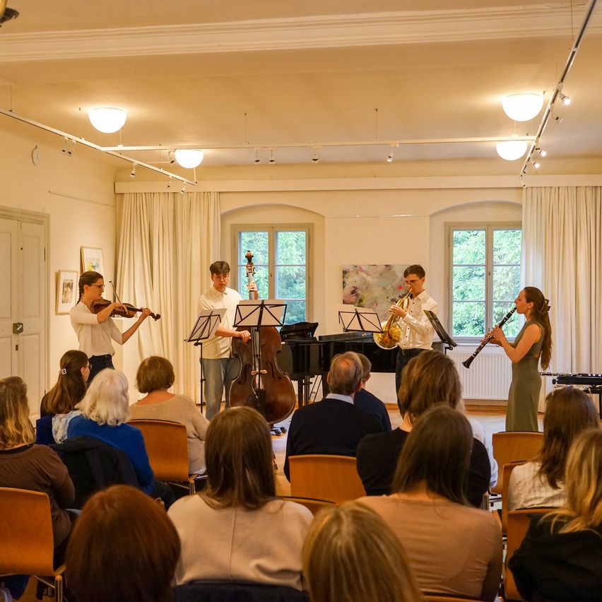 Ein Quartett von Musikern tritt auf der Bühne in einem gut beleuchteten Raum auf, vor ihnen ein Publikum. Die Musiker spielen Violine, Cello, Trompete und Klarinette.