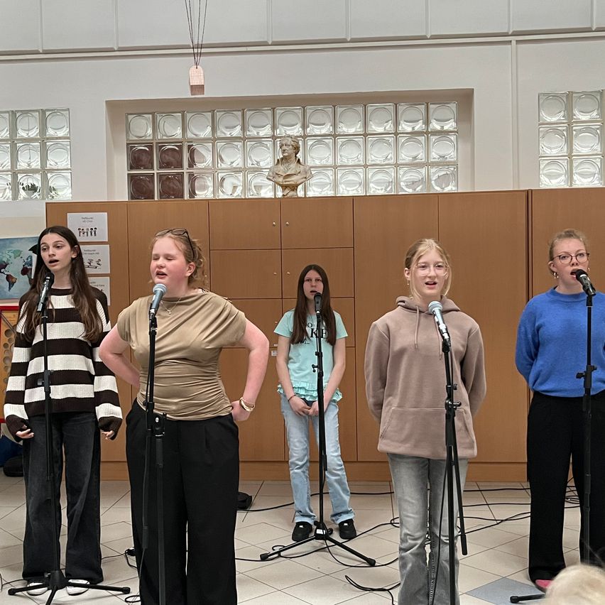 Fünf Frauen stehen vor Mikrofonen und singen in einer Halle mit gefliesten Böden und einer hölzernen Wand dahinter.