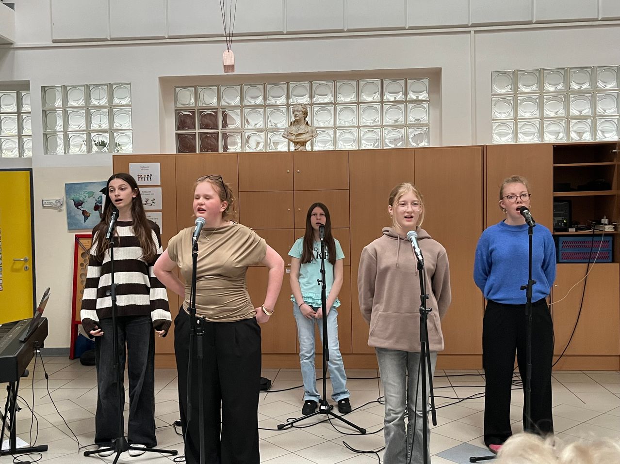 Fünf Frauen stehen vor Mikrofonen und singen in einer Halle mit gefliesten Böden und einer hölzernen Wand dahinter.