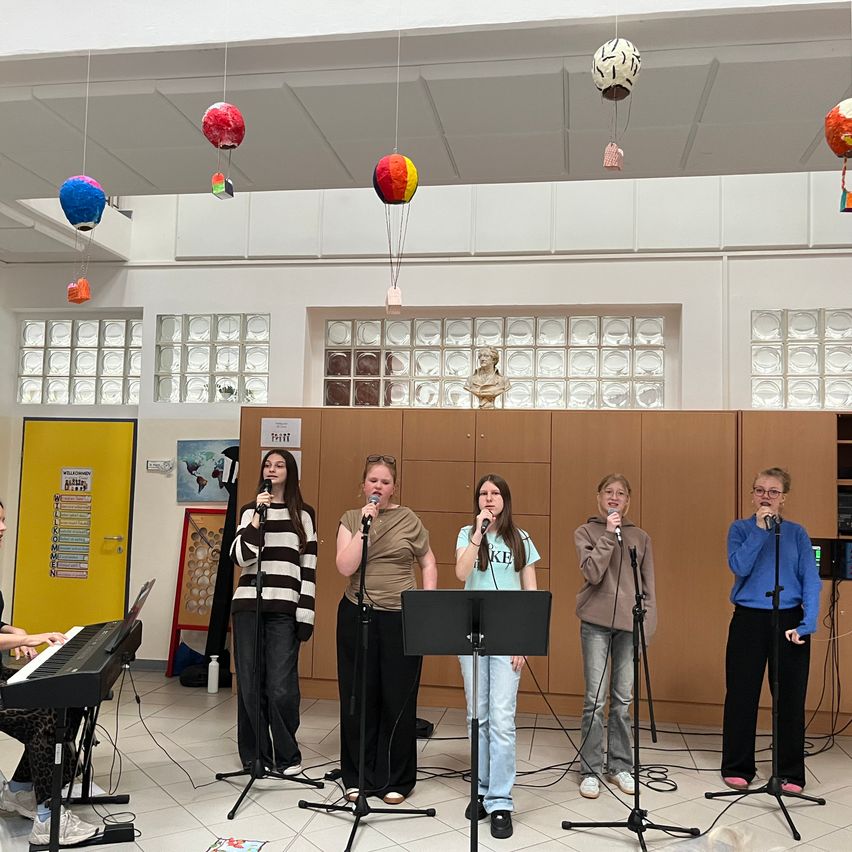 Fünf Frauen singen in Mikrofone auf einer Bühne mit einem Pianisten. Bunte Luftballons hängen von der Decke. Hinter den Frauen befinden sich Holzschränke und eine Glaswand.