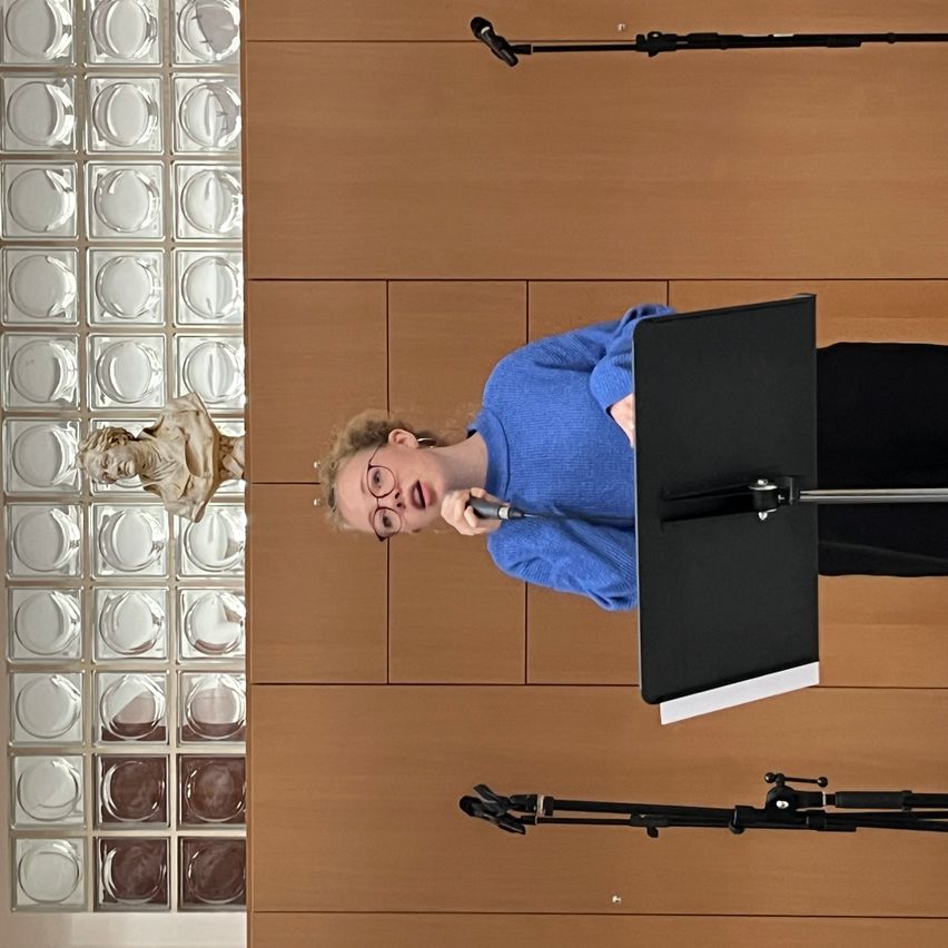Eine Frau in einem blauen Pullover und mit Brille singt in ein Mikrofon in einem Raum mit Holzpaneelen und einer Glasmosaikwand.