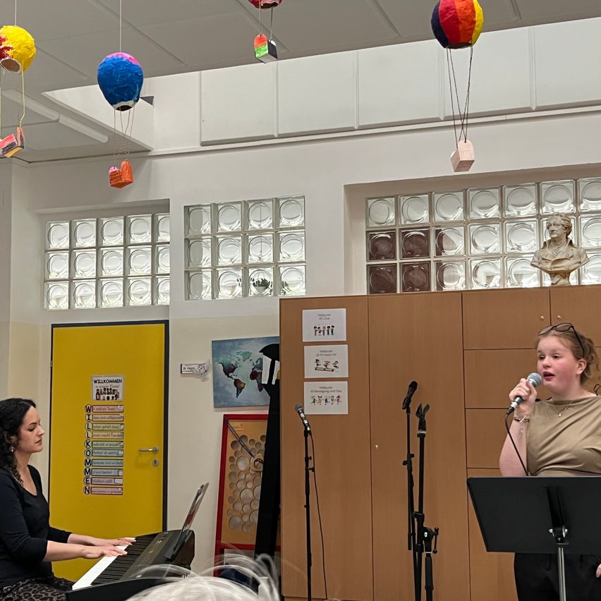 Zwei Frauen singen ein Lied in einem Klassenzimmer. Eine singt, während die andere Klavier spielt. Bunte Papierlaternen hängen von der Decke.