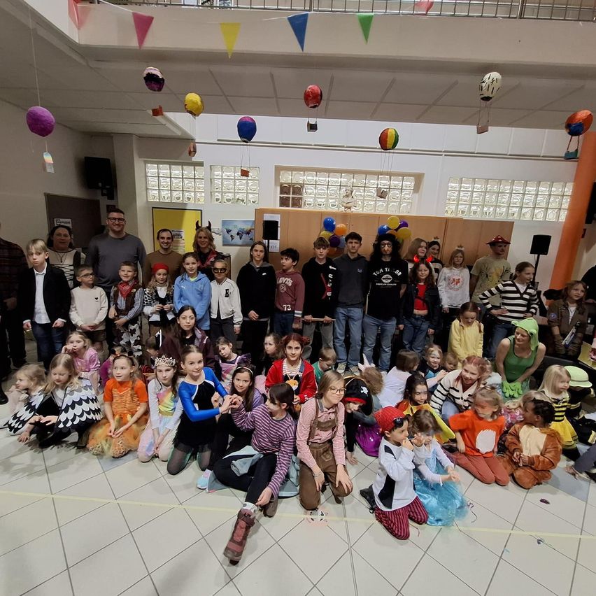 Eine Gruppe von Menschen, darunter Kinder in Kostümen, versammelt sich in einem Saal für ein Foto. Bunte Luftballons hängen von der Decke ab.