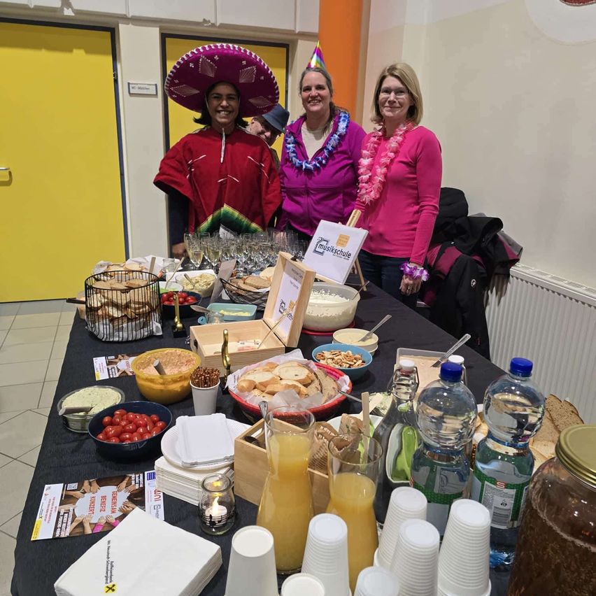 Drei Frauen stehen hinter einem Tisch mit verschiedenen Speisen und Getränken, eine trägt einen Sombrero, eine andere ein rosa Shirt und die dritte ein lila Shirt. Ein Whiteboard mit Schrift steht auf dem Tisch.