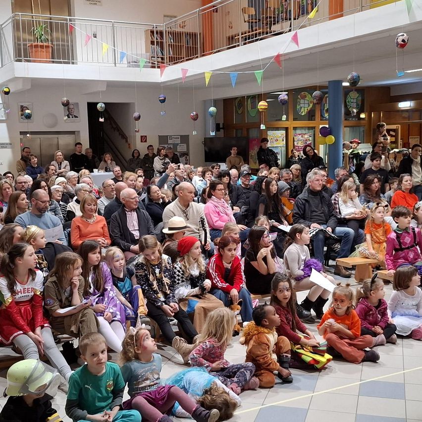 Eine große Gruppe von Menschen und Kindern sitzt auf dem Boden in einer Halle, einige schauen zu, während andere Papiere halten. Bunte Dekorationen hängen von der Decke.