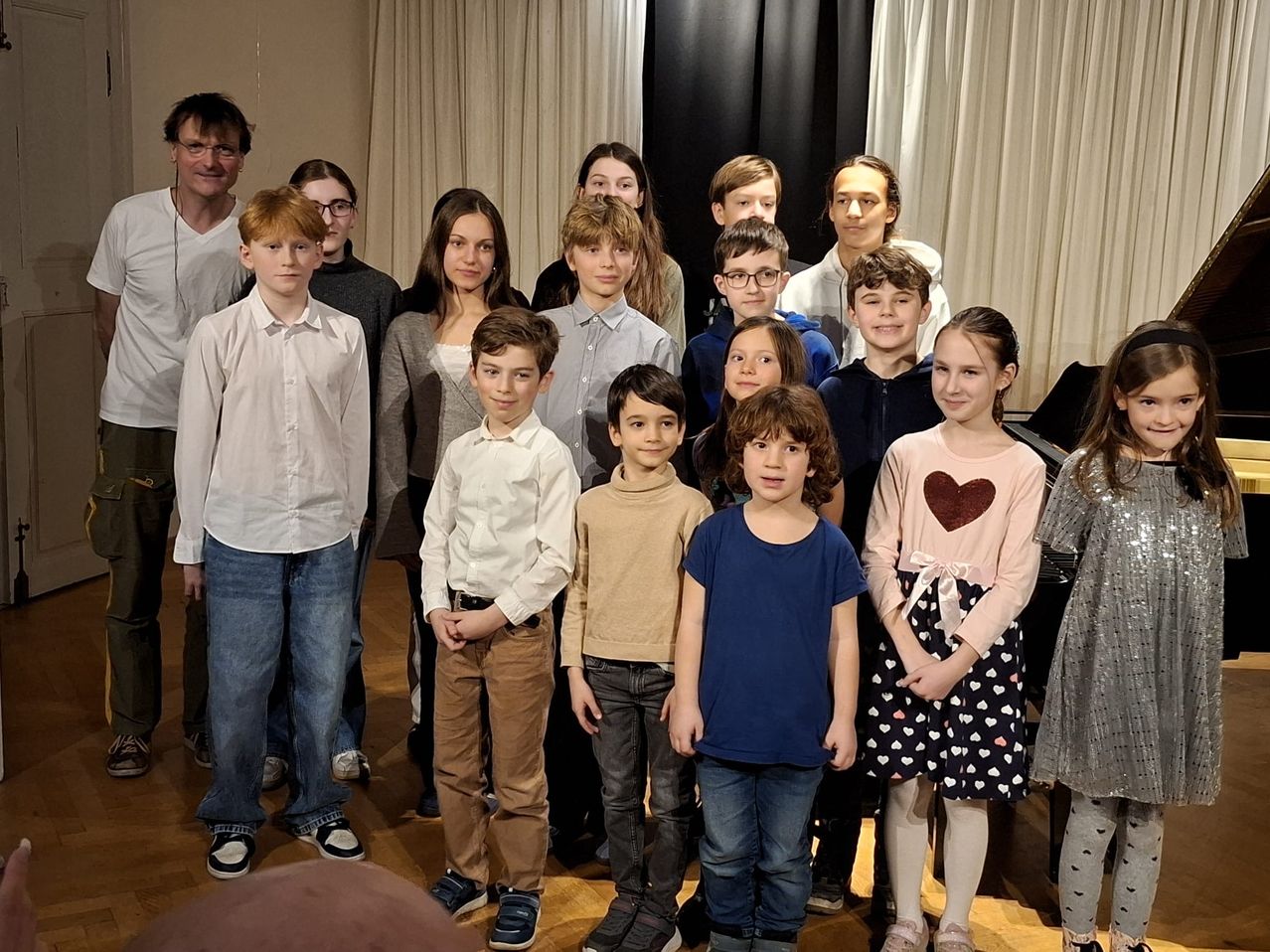 Eine Gruppe von Kindern und Erwachsenen steht in einem Raum mit weißen Vorhängen und lächelt für ein Foto. Die Kinder sind in Freizeitkleidung gekleidet. Ein erwachsener Mann mit Brille steht links, und ein Klavier ist hinter der Gruppe zu sehen.