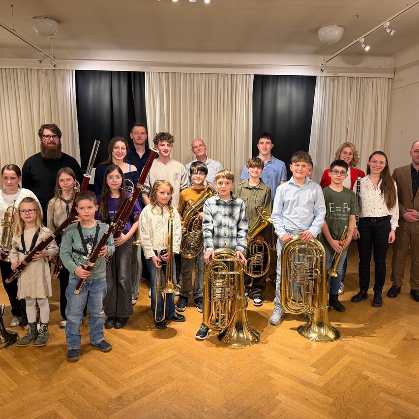 Eine Gruppe von Musikern, darunter Kinder und Erwachsene, posiert für ein Foto, während sie verschiedene Blechblasinstrumente halten. Sie stehen in einem Raum mit Holzboden.