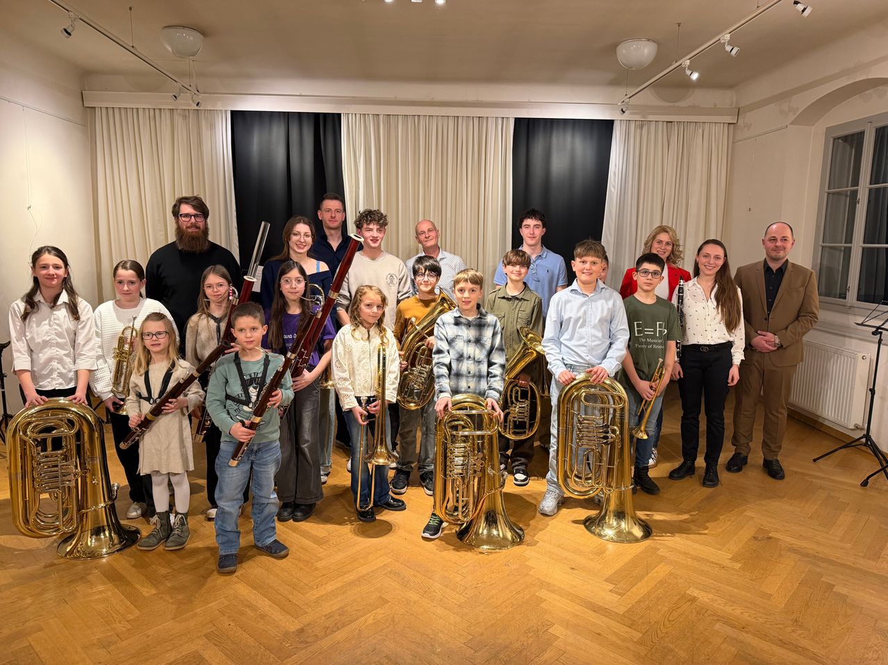 Eine Gruppe von Musikern, darunter Kinder und Erwachsene, posiert für ein Foto, während sie verschiedene Blechblasinstrumente halten. Sie stehen in einem Raum mit Holzboden.