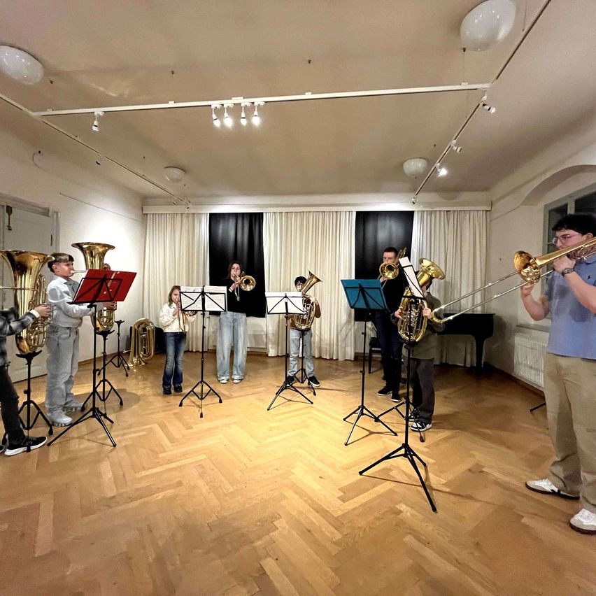 Eine Gruppe von Musikern übt in einem gut beleuchteten Raum mit Holzböden und hängenden Lampen. Sie spielen verschiedene Blechblasinstrumente, einige stehend und andere sitzend. Musikblätter stehen auf Ständern, und ein Klavier ist in der Nähe.