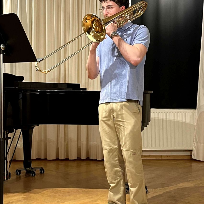 Ein Mann steht in einem Raum mit Klavier, Heizkörper und weißen Vorhängen und spielt Posaune.