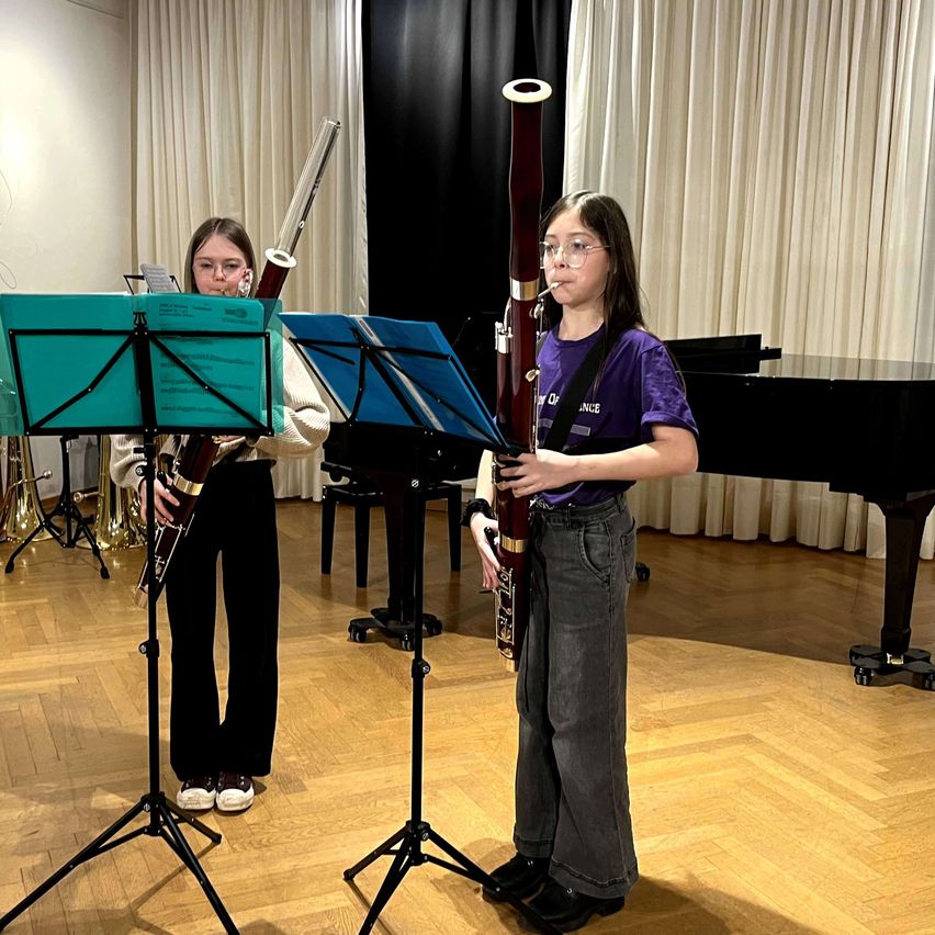 Zwei Mädchen spielen das Fagott in einem Raum mit einem Klavier und Notenständern im Hintergrund.