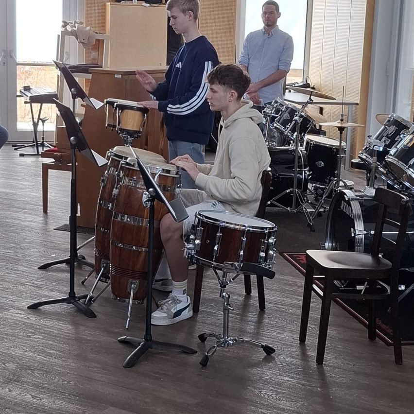 Drei junge Männer spielen in einem Raum Schlagzeug. Einer von ihnen sitzt auf einem Hocker. Es gibt andere Trommeln und Becken um sie herum.