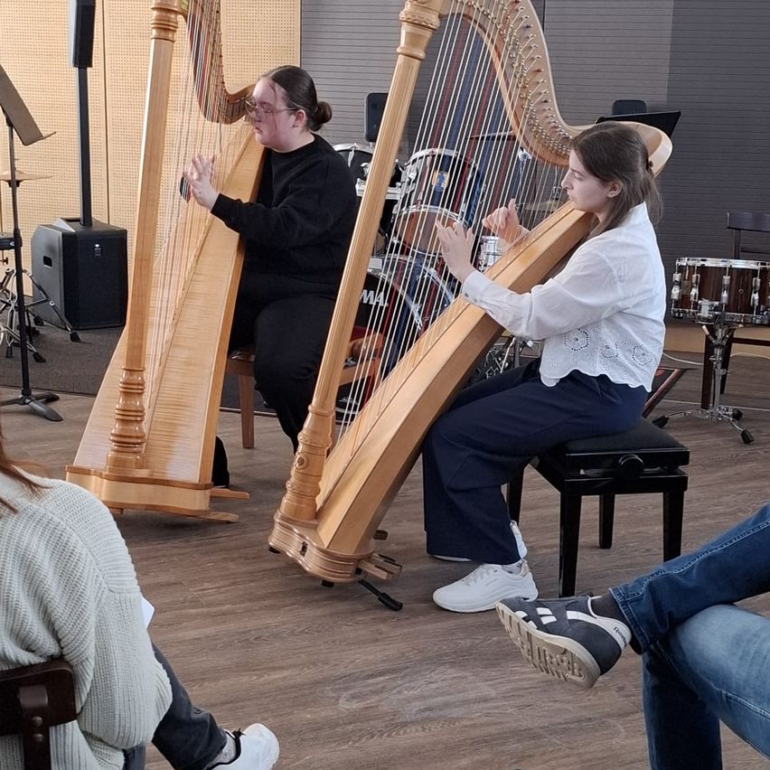 Zwei Frauen spielen Harfen in einem Raum mit Holzboden, mit Trommeln und einem Lautsprecher in der Nähe. Eine Frau sitzt auf einem Stuhl, während eine andere steht.