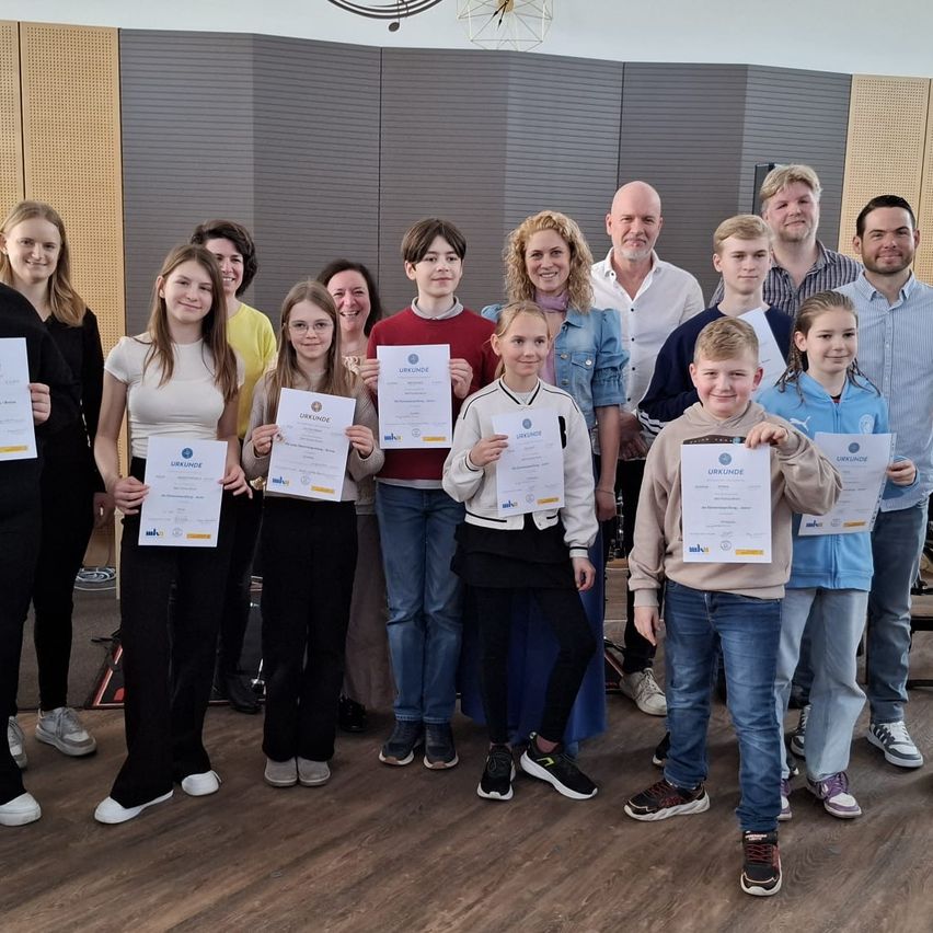 Eine Gruppe von Menschen steht zusammen und hält Urkunden in der Hand. Sie lächeln und stehen in einem Raum mit Holzboden und einer Wand dahinter. Einige tragen Turnschuhe, und eine Person trägt eine Jacke. Sie scheinen eine Errungenschaft zu feiern.