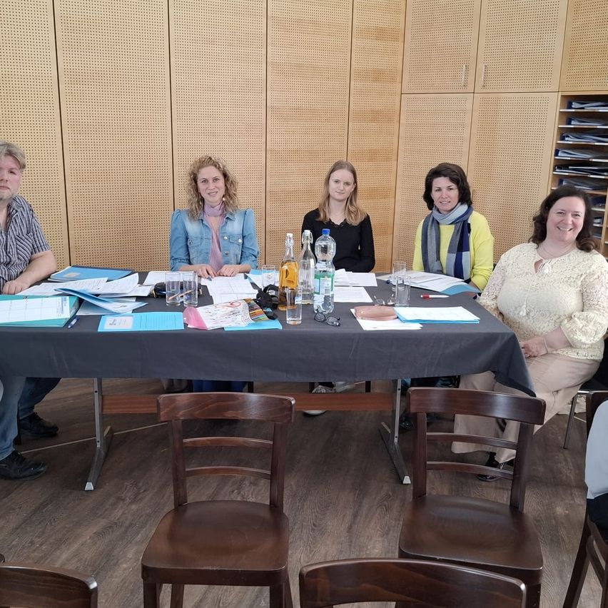 Sechs Personen sitzen um einen Tisch in einem Konferenzraum. Sie haben Papiere, Gläser und Flaschen auf dem Tisch. Dahinter ist eine Wand mit Regalen.