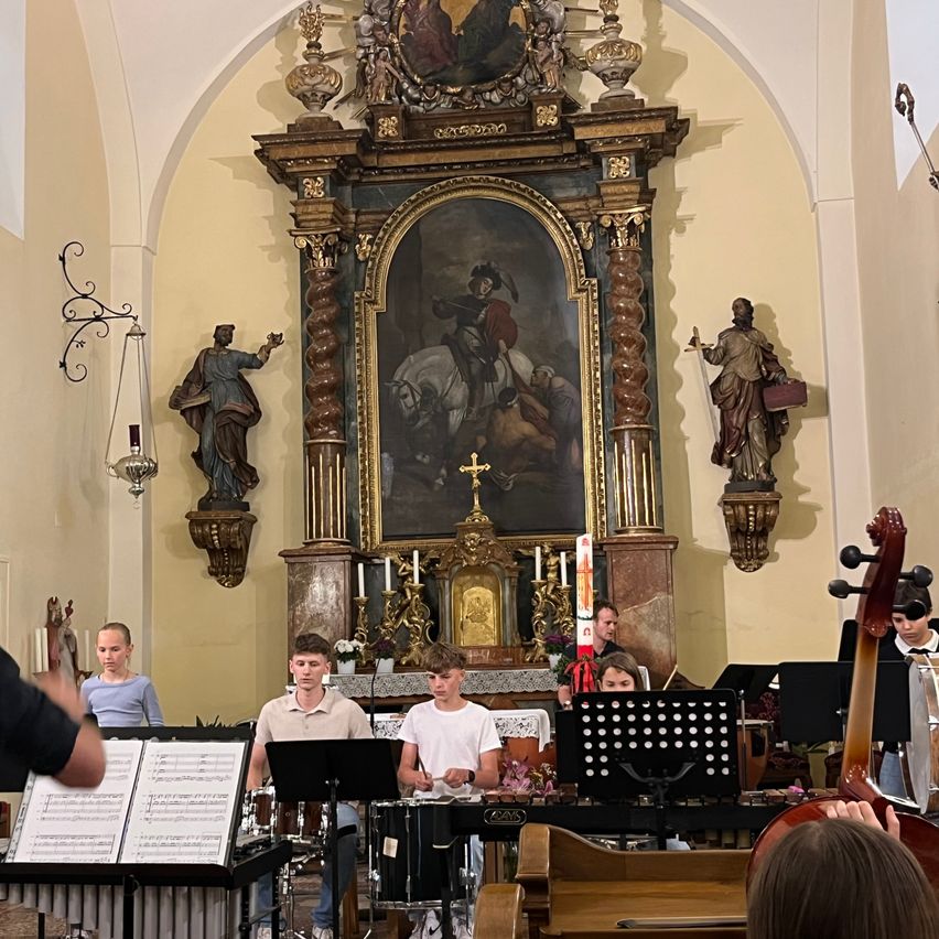 Ein kleines Orchester spielt in einer Kirche mit mehreren Zuschauern. Die Instrumente umfassen Trommeln und ein Xylophon.