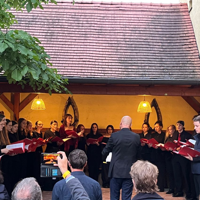 Ein Chor aus Frauen und Männern, gekleidet in Schwarz, singt in einem Gebäude mit einem roten Dach. Ein Dirigent führt sie an, und einige Leute sehen von unten zu.