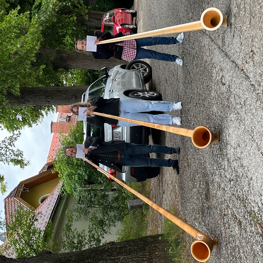 Drei Personen, die lange Holzhörner halten, stehen neben einem geparkten Auto auf einer Kiesstraße, mit Bäumen und einem Haus im Hintergrund.