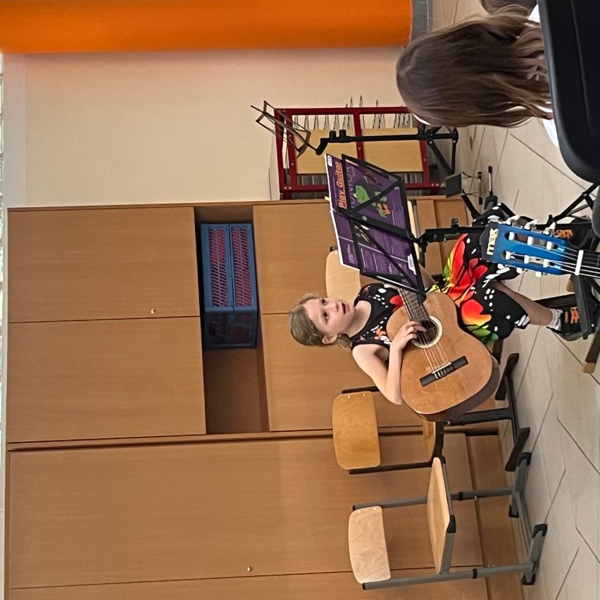 Ein junges Mädchen spielt Gitarre in einem Klassenzimmer. Sie sitzt, mit einem Notenständer davor. Hinter ihr ist eine Aufbewahrungsbox zu sehen, und mehrere Stühle und ein Schrank sind aufgestellt.