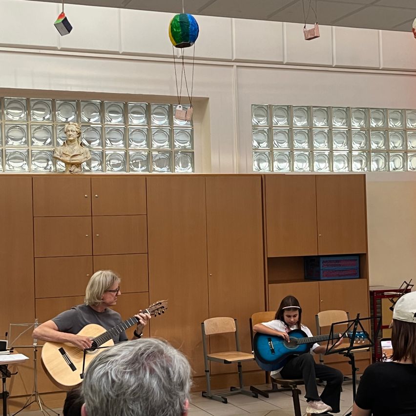 Zwei Personen spielen Gitarren in einem Raum mit einem hölzernen Schrank und einer Glaswand hinter ihnen. Ein Brustbild und bunte Luftballons sind über der Wand.