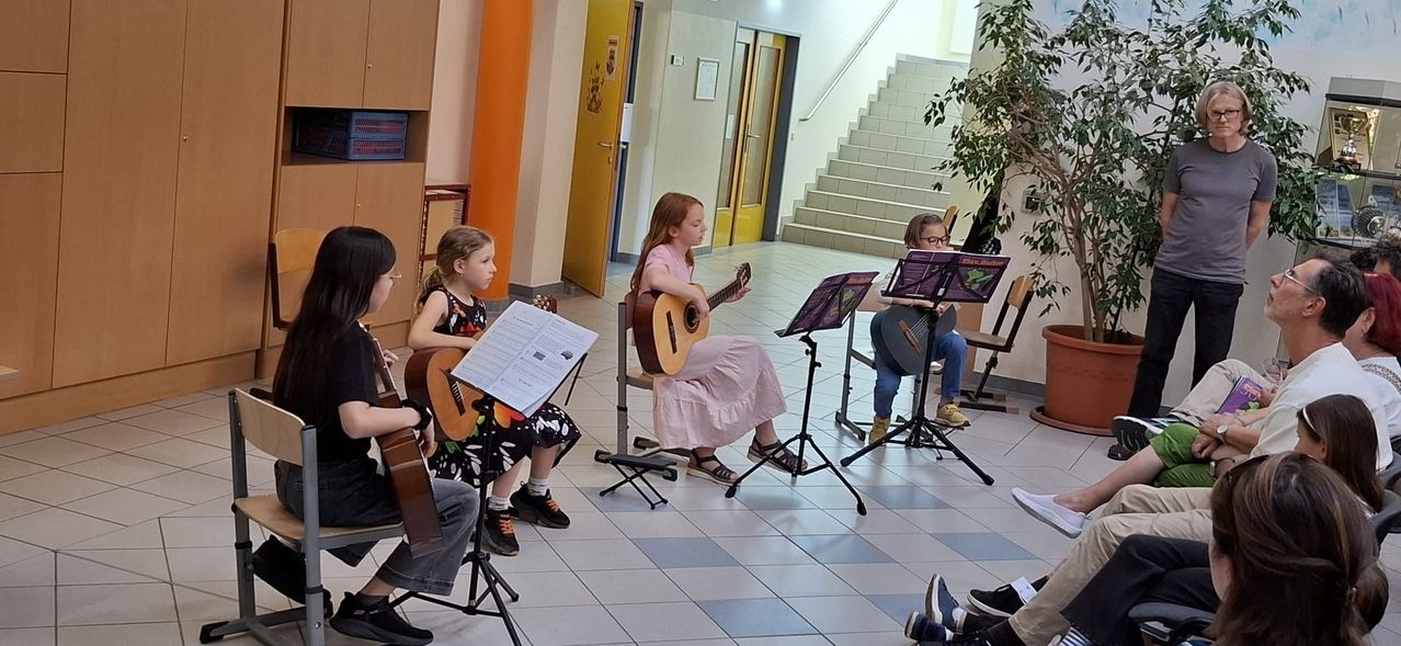 Vier Mädchen in einem Flur, jede spielt eine Gitarre. Sie sitzen auf Stühlen und haben Notenständer vor sich. Dahinter befindet sich eine Treppe.