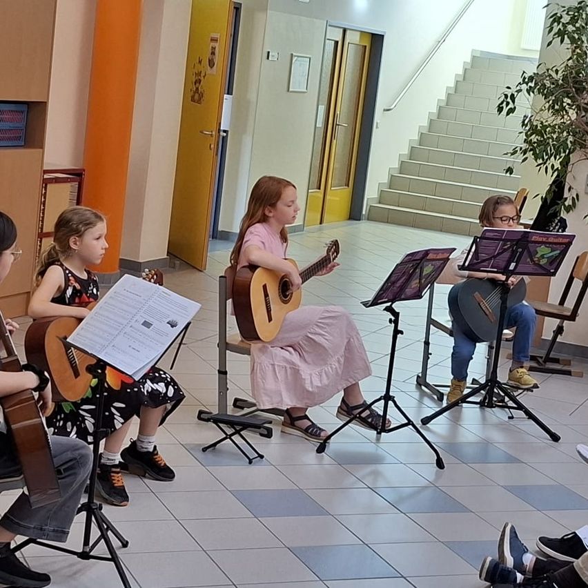 Vier Mädchen in einem Flur, jede spielt eine Gitarre. Sie sitzen auf Stühlen und haben Notenständer vor sich. Dahinter befindet sich eine Treppe.