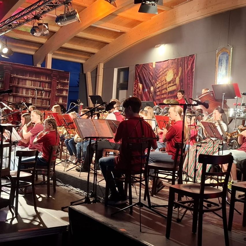 Ein Orchester steht auf der Bühne in einem Raum mit Holzbalken, Bücherregalen und Bannern. Musiker sitzen auf Stühlen mit Notenständern und spielen verschiedene Instrumente. Scheinwerfer und Lampen beleuchten den Raum.