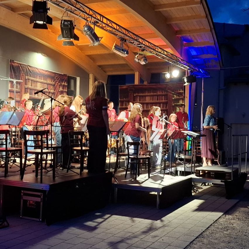 Eine Band spielt auf einer Bühne mit verschiedenen Instrumenten unter blauen Lichtern bei Nacht. Eine Frau steht vorne, und Menschen stehen hinter ihr.