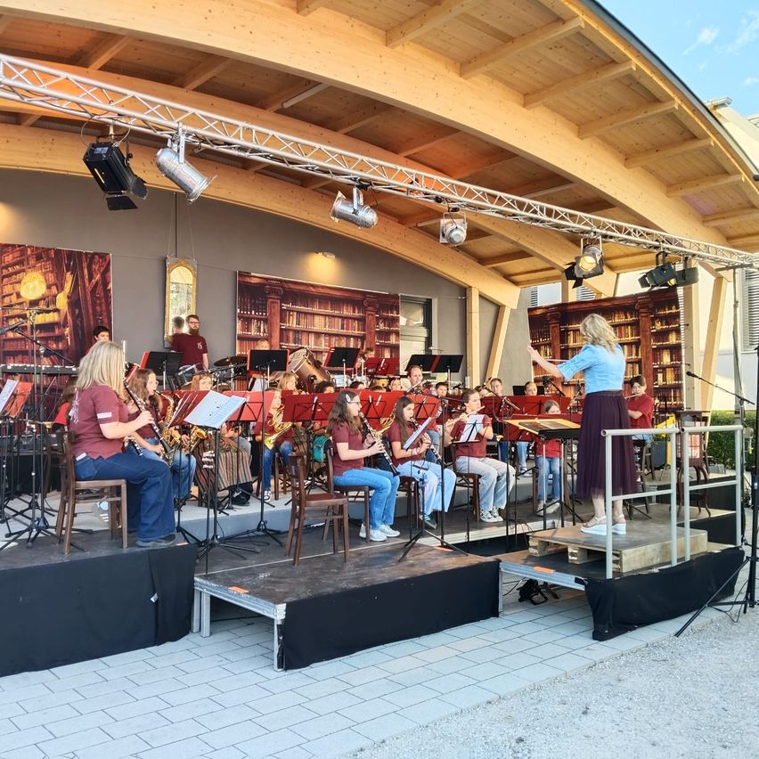 Eine Band spielt auf der Bühne mit einer Frau davor. Einige spielen Blasinstrumente. Es gibt Lichter und ein Mikrofon auf der Bühne.
