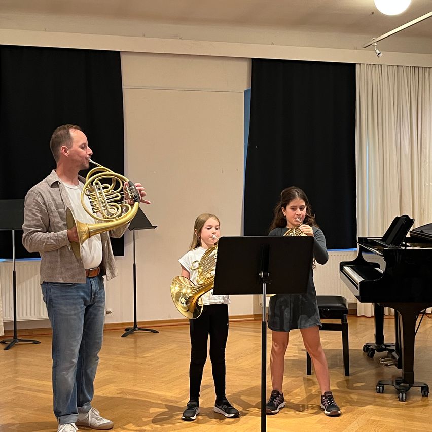 Ein Mann und zwei Mädchen üben gemeinsam in einem Raum mit Holzböden Musik. Der Mann spielt das Horn, während die Mädchen Trompete und Horn spielen. Hinter ihnen steht ein schwarzer Klavier.