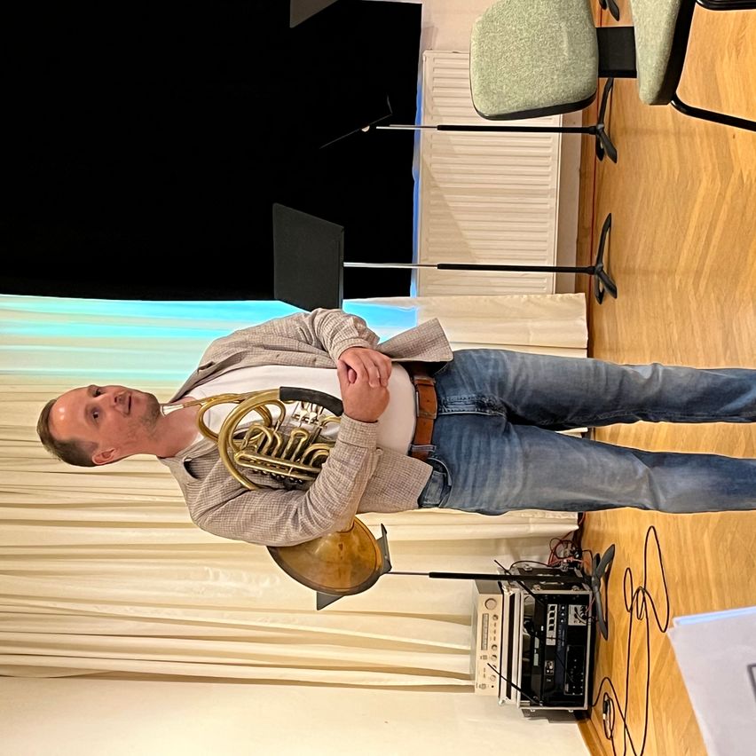 Ein Mann hält ein Waldhorn in der Hand und steht auf der Bühne, er trägt eine leichte Jacke und Jeans. Hinter ihm befindet sich ein Mikrofonstativ und ein Lautsprecher.