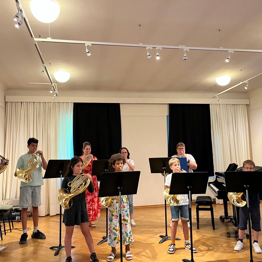 Eine Gruppe von Kindern und Erwachsenen spielt Blechblasinstrumente in einem Raum. Sie stehen in einer Reihe, mit einem Klavier und Notenständern im Hintergrund.