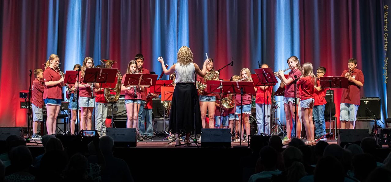 Eine Band tritt auf der Bühne auf, spielt Instrumente wie Trompeten und Posaunen und wird von einer Frau dirigiert. Die Kinder stehen hinter ihren Notenständern.