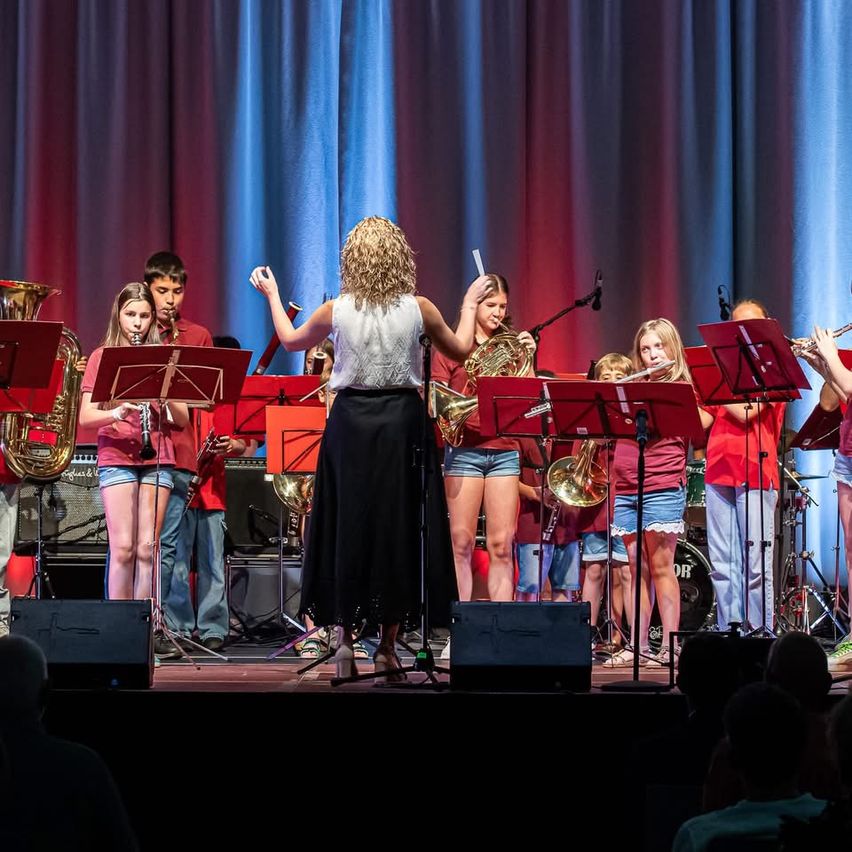 Eine Band tritt auf der Bühne auf, spielt Instrumente wie Trompeten und Posaunen und wird von einer Frau dirigiert. Die Kinder stehen hinter ihren Notenständern.