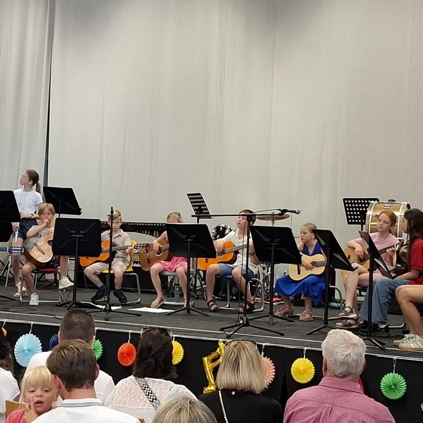 Eine Gruppe von Kindern spielt Gitarren auf der Bühne mit Notenständern und Trommeln, während Erwachsene und Kinder von unten zuschauen.