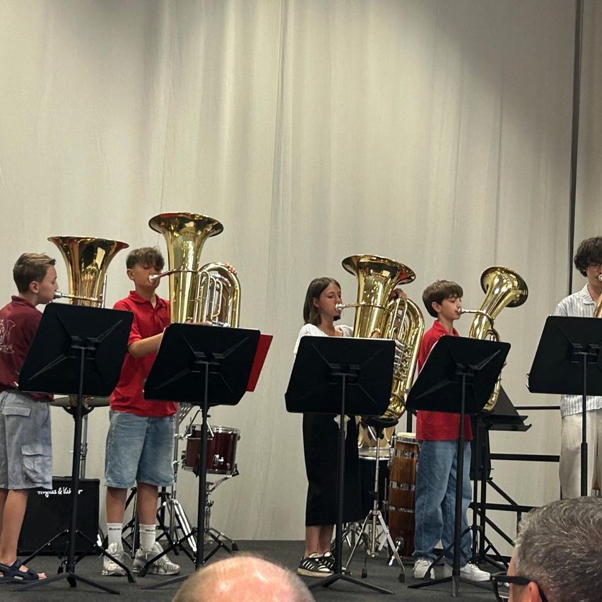 Eine Gruppe von Kindern spielt Tuba auf der Bühne mit Trommeln im Hintergrund. Sie tragen rote Hemden und spielen für ein Publikum.