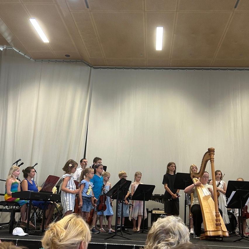 Eine Gruppe von Kindern und Erwachsenen tritt auf einer Bühne auf. Sie spielen Musikinstrumente, darunter eine Harfe und Violinen. Die Bühne hat weiße Vorhänge und Scheinwerfer.