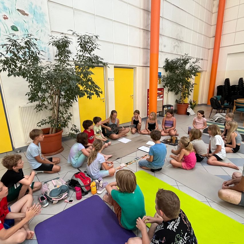 Eine Gruppe von Kindern sitzt auf dem Boden in einem Raum mit gelben Türen und Topfpflanzen, einige spielen Gitarre, andere schauen zu.