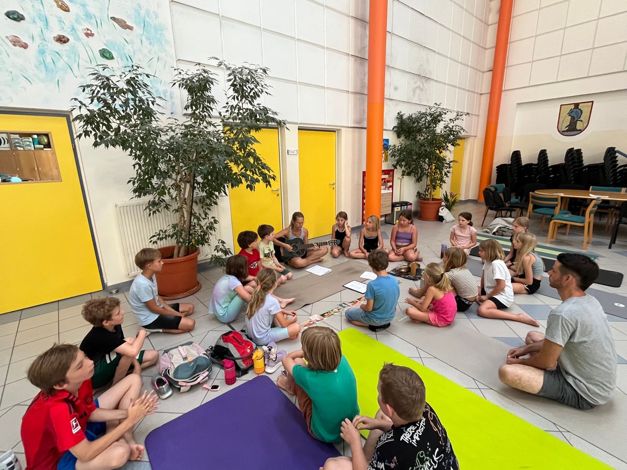 Eine Gruppe von Kindern sitzt auf dem Boden in einem Raum mit gelben Türen und Topfpflanzen, einige spielen Gitarre, andere schauen zu.