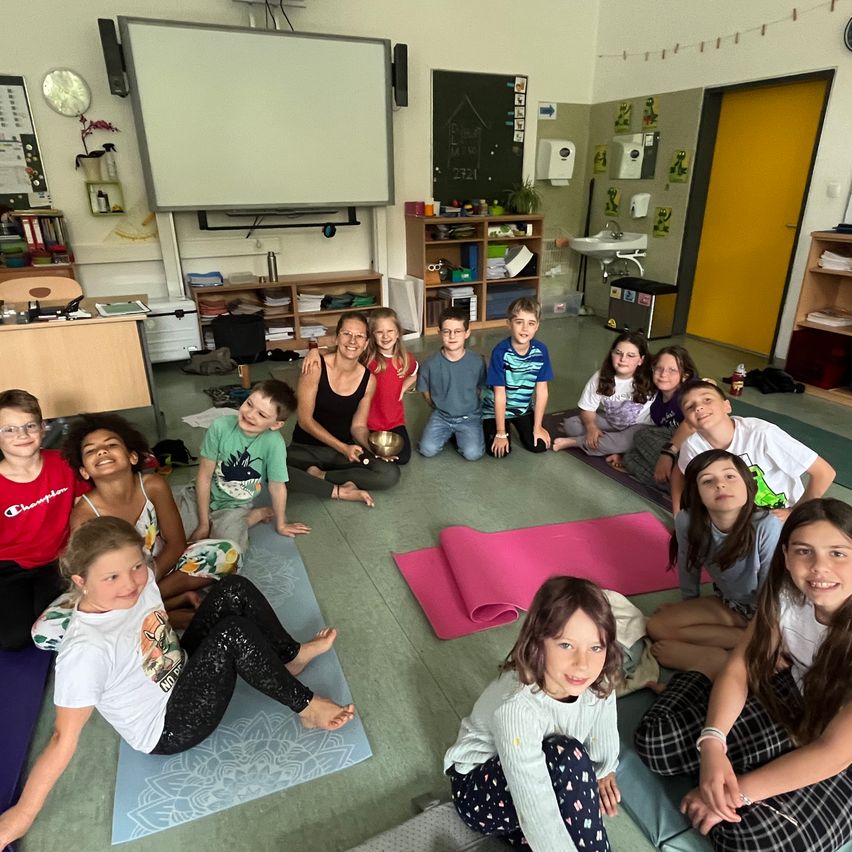 Eine Gruppe von Kindern und einer Frau praktiziert Yoga in einem Raum. Einige sitzen im Schneidersitz auf Yogamatten, andere auf Stühlen, während rosa Matten und Yoga-Zubehör auf dem Boden verstreut sind. Eine Tafel und Regale mit verschiedenen Gegenständen sind im Hintergrund zu sehen.
