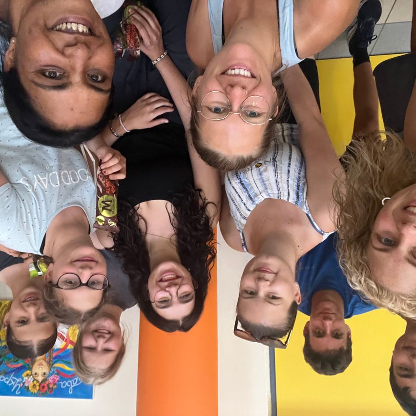 Eine Gruppe lächelnder Personen, einige mit Brillen, posiert für ein Selfie in einem farbenfrohen Raum. Ein Poster ist an der Wand hinter ihnen sichtbar.