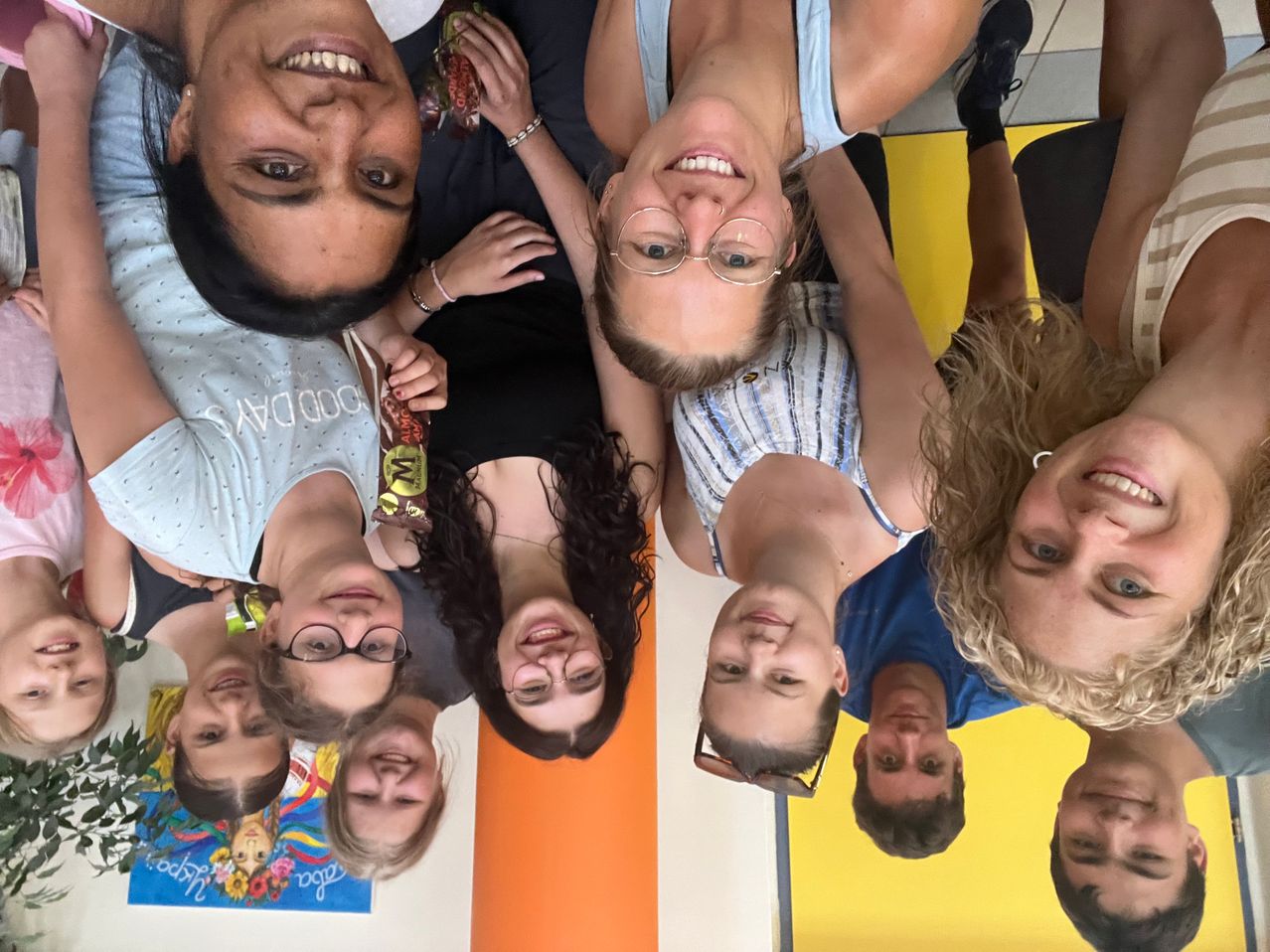 Eine Gruppe lächelnder Personen, einige mit Brillen, posiert für ein Selfie in einem farbenfrohen Raum. Ein Poster ist an der Wand hinter ihnen sichtbar.
