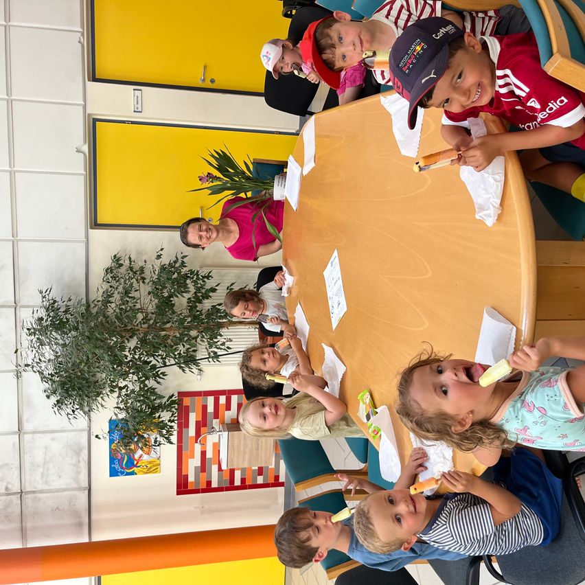 Eine Gruppe von Kindern und Erwachsenen sitzt um einen Tisch und isst Eis in einem Raum mit gelben Türen.