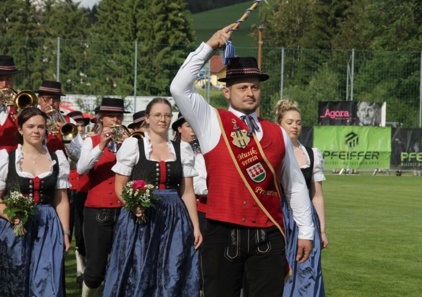Eine Gruppe von Menschen in traditionellen Kostümen geht auf einem Feld. Ein Mann hält einen Stab und schaut nach vorne. Eine Frau hält Blumen und geht neben ihm. Hinter ihnen spielen andere Instrumente. Ein Zaun und Bäume sind im Hintergrund.