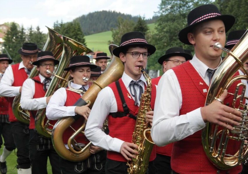 Eine Blaskapelle in traditioneller bayerischer Tracht, einschließlich roter Westen und schwarzer Hüte, steht auf einer Rasenfläche und spielt Instrumente wie Tubas, Saxophone und Trompeten.