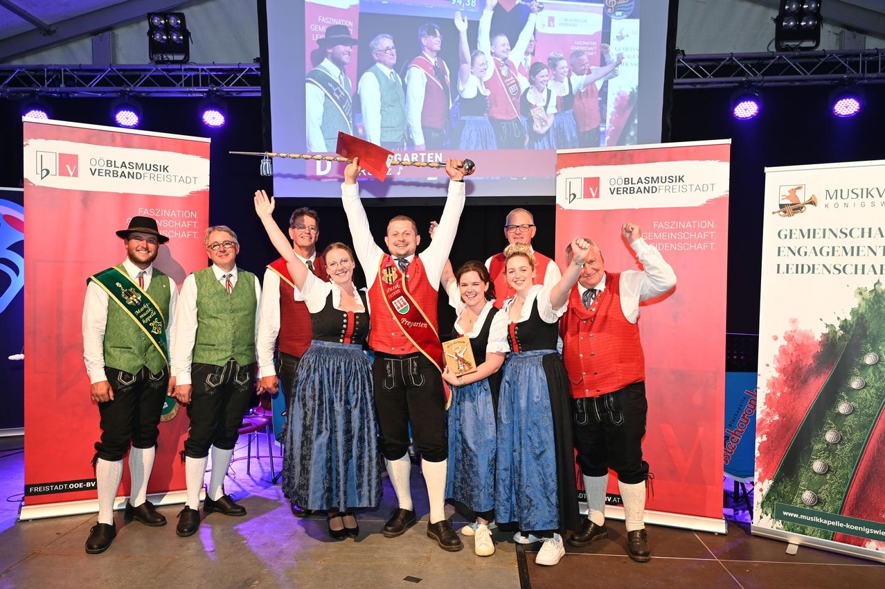 Eine Gruppe von Menschen in traditionellen Kostümen steht auf der Bühne und feiert mit erhobenen Armen. Ein Mann in der Mitte hält ein rotes Banner und eine Trophäe. Hinter ihnen ist eine große Leinwand, die Menschen in traditionellen Kostümen zeigt.