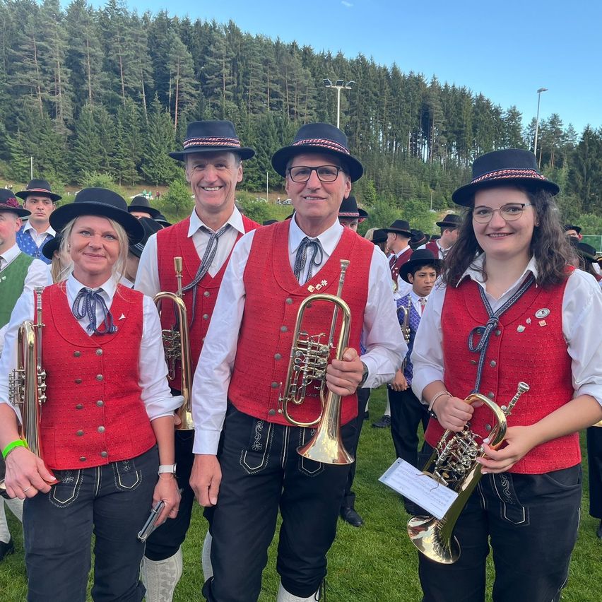 Eine Gruppe von Musikern in traditioneller Kleidung steht auf einem Rasenfeld, hält Musikinstrumente in den Händen und hat Bäume und blauen Himmel im Hintergrund.