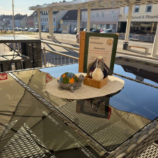 Ein Glastisch hat eine kleine Schildkrötenfigur und eine Menükarte. Dahinter ist ein Gebäude mit dem Schild 'Restaurant & Pizzeria' zu sehen. Das Wetter ist sonnig.